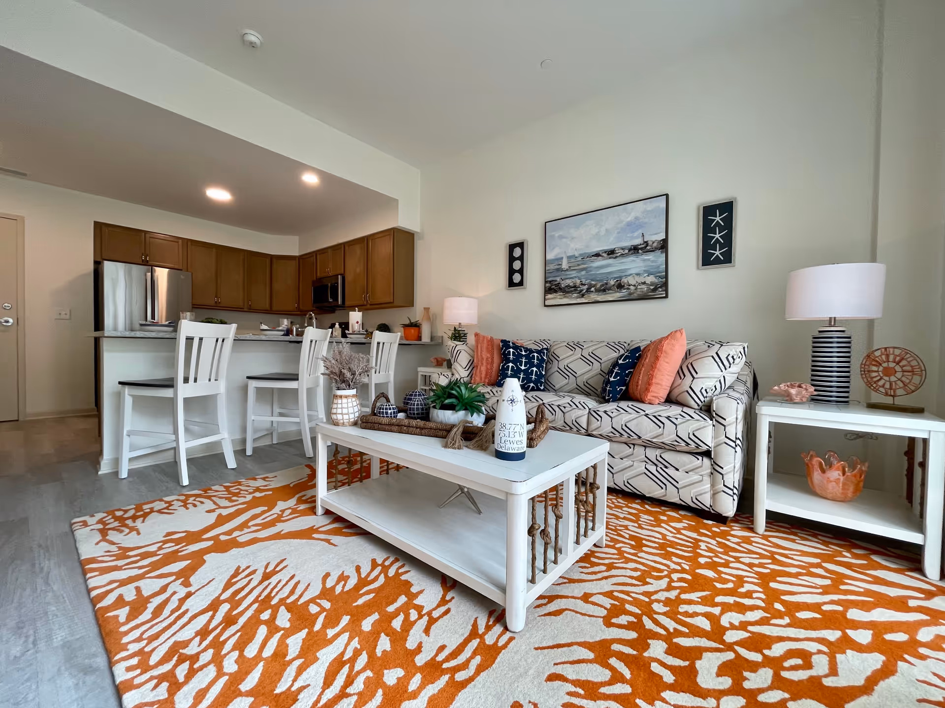 A cozy living room and kitchen area with a patterned beige sofa adorned with coral and navy blue anchor-themed pillows. A white coffee table with decorative items sits on an orange and white patterned rug. The kitchen features wooden cabinets, a stainless steel refrigerator, and a breakfast bar with three white chairs. The walls are decorated with coastal-themed artwork and the room is well-lit with ceiling lights and table lamps.