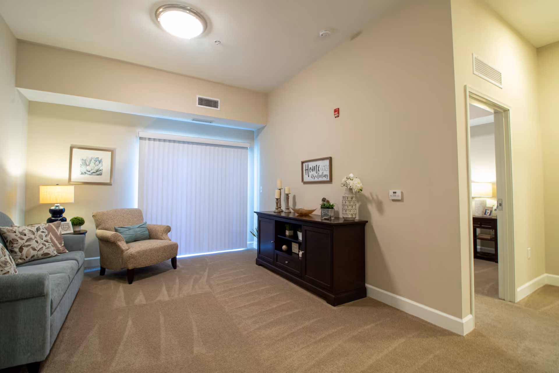 A cozy living room area with beige carpet and walls, featuring a blue sofa with patterned pillows, a beige armchair, a side table with a lamp and small plant, a dark wooden cabinet decorated with candles, plants, and a vase with flowers. A framed sign on the wall reads 'Home is not a place, it's a feeling.' Vertical blinds cover a large window or sliding door, and an open doorway leads to another room with a lamp and furniture visible.