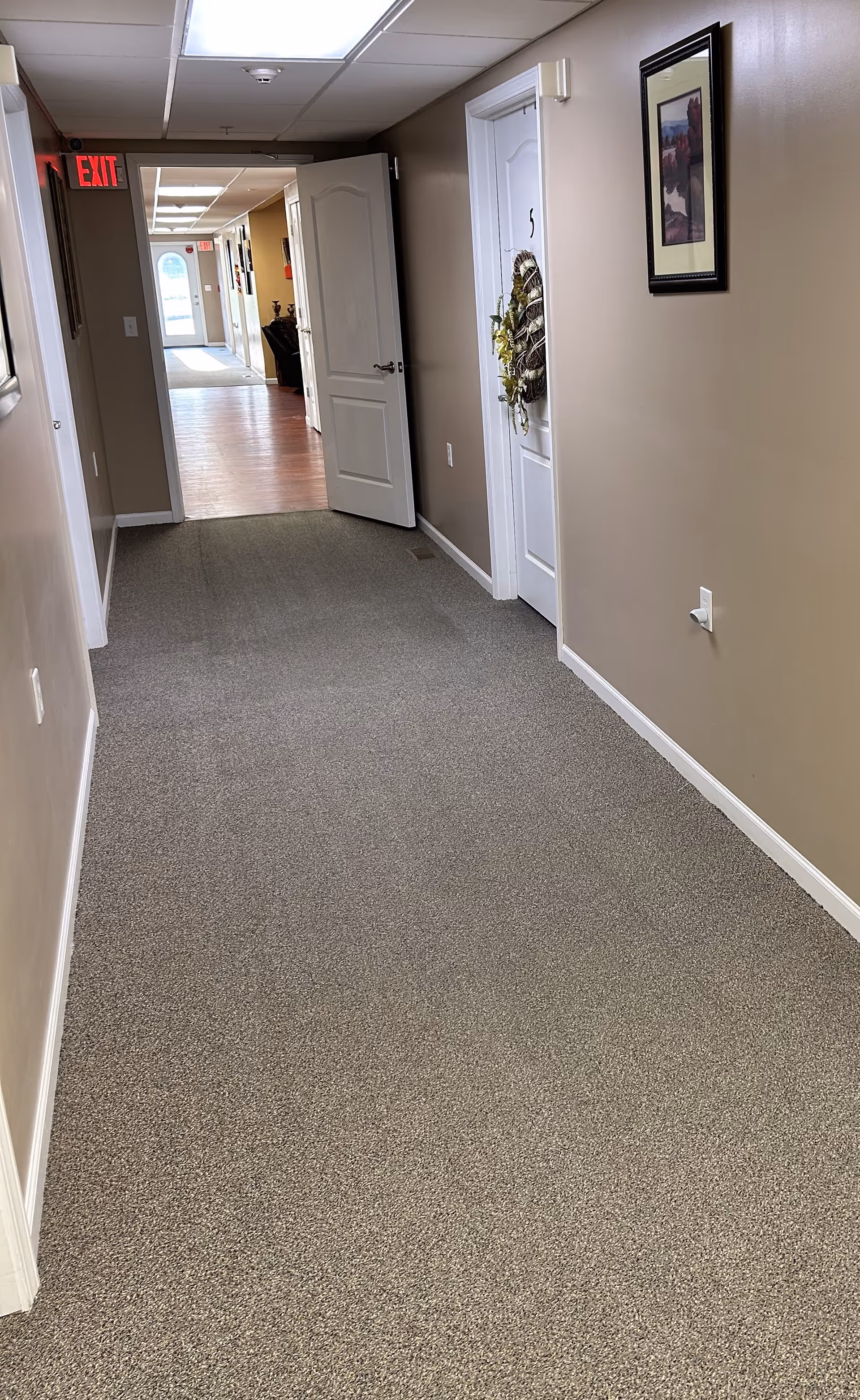 Carpeted hallway in a residential care facility with beige walls, several doors (one decorated with a wreath) and an exit sign at the far end.
