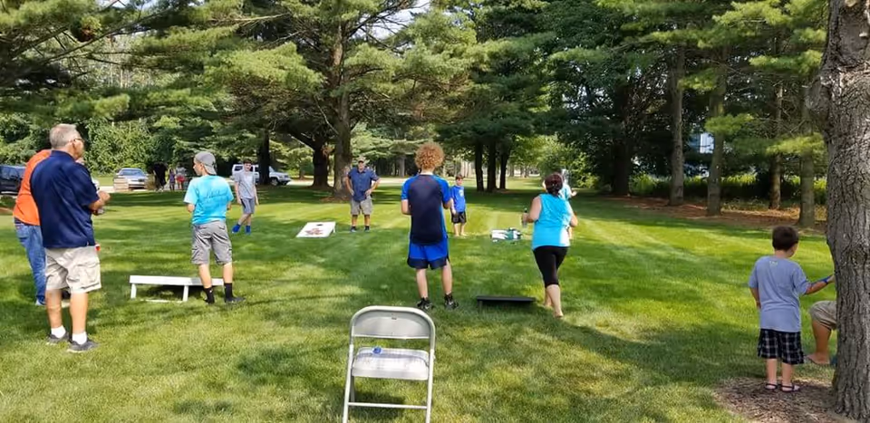 A group of people, including children and adults, playing cornhole on a grassy area surrounded by trees on a sunny day. Several cornhole boards are set up on the lawn, and a folding chair is visible in the foreground.