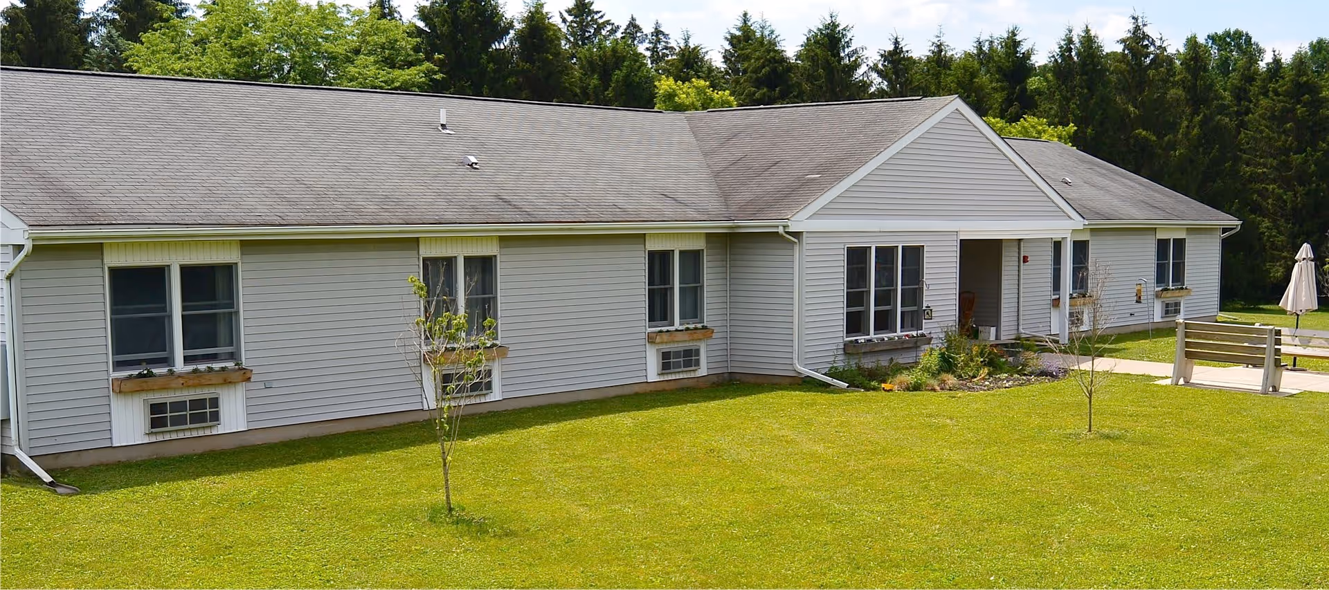 Single-story building with light gray siding and a dark shingled roof, surrounded by green grass and trees in the background. There are several windows with flower boxes and a small covered entrance. A wooden bench and a closed umbrella are visible on the right side of the image.