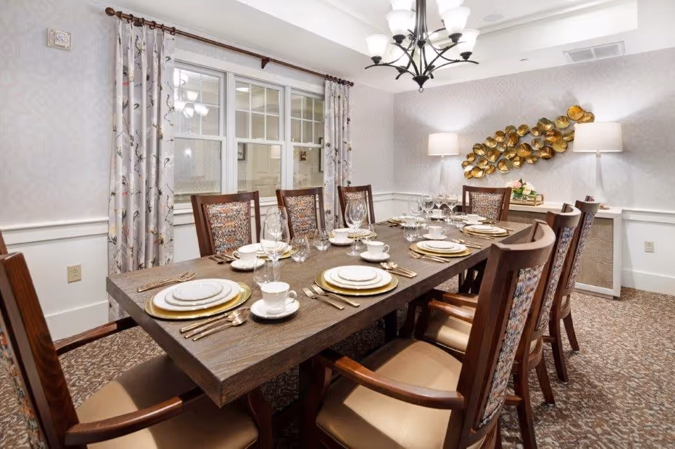 A formal dining room with a long wooden table set for eight people. The table is arranged with white plates, gold chargers, cups, wine glasses, and silverware. The room features patterned carpet, floral curtains on the window, a chandelier overhead, two floor lamps, and a decorative gold wall art piece above a sideboard.