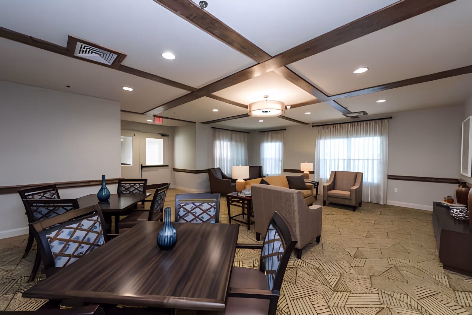 A well-lit common area in a senior living facility featuring multiple dark wood tables with chairs, each table decorated with a blue vase. The room has beige carpet with a geometric pattern, beige walls with dark wood trim, and large windows with sheer white curtains. There are several upholstered armchairs and sofas arranged around side tables with lamps, and the ceiling has recessed lighting and wooden beams.