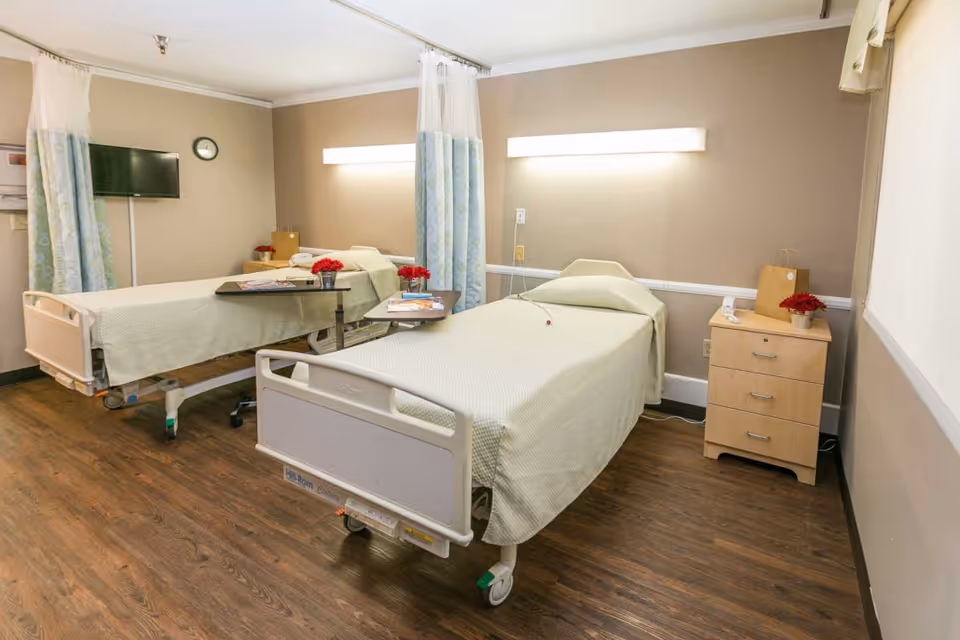 A room with two hospital-style beds, each covered with light green bedspreads. Each bed has a small overbed table with magazines and a red flower vase. There are privacy curtains between the beds, a wall-mounted TV, a clock, and a wooden nightstand with drawers and a red flower vase. The room has wood flooring and beige walls with white trim.