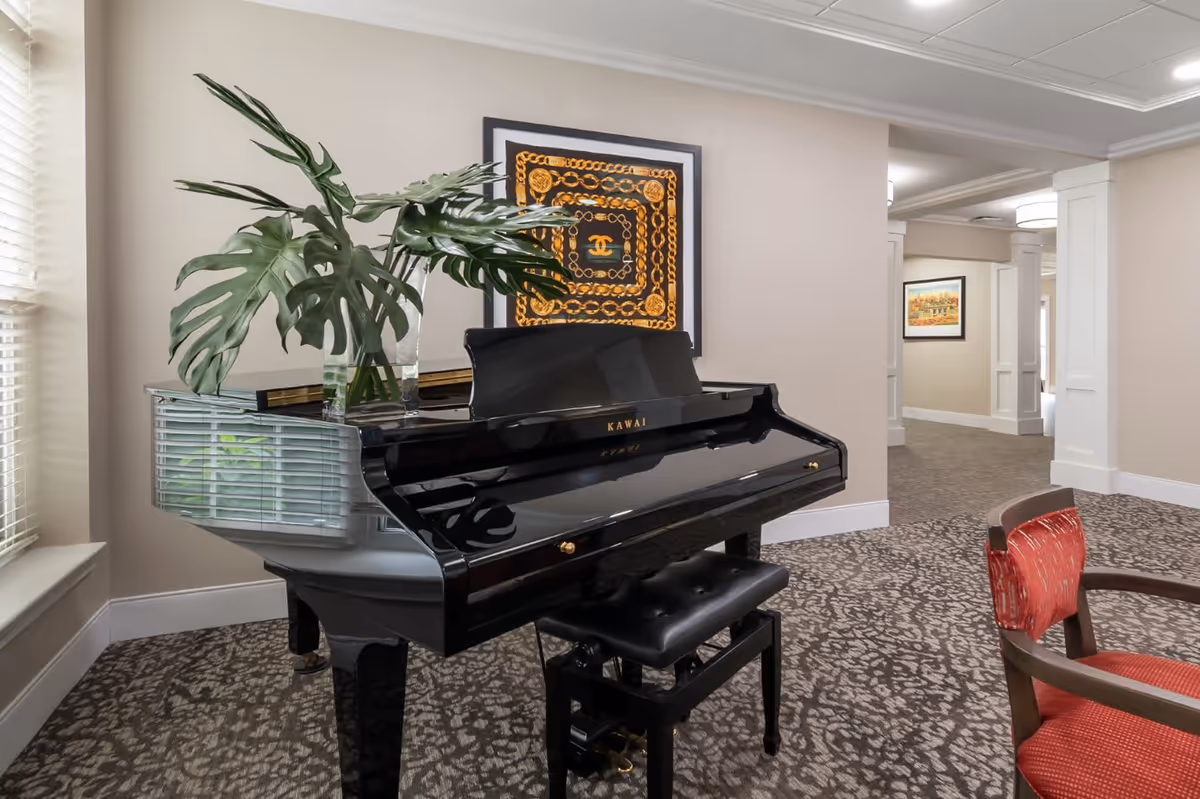 Interior view of a room featuring a black Kawai grand piano with a vase of large green leaves on top. Behind the piano is a framed artwork with a gold chain pattern on the wall. To the right, there is a red cushioned chair and a hallway with beige walls and carpeted floors.
