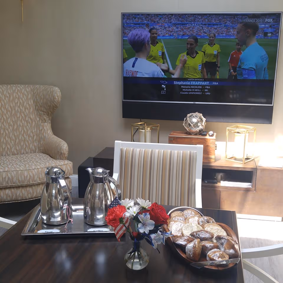 A cozy living room area with a beige patterned armchair, a striped chair, and a dark wooden table. On the table, there is a tray with two silver coffee pots labeled Decaf and Regular, a small vase with red, white, and blue flowers and a miniature American flag, and a wooden bowl filled with assorted wrapped pastries. A flat-screen TV mounted on the wall displays a soccer match with referees and players. The room is softly lit with natural light and decorative lanterns on a wooden cabinet beneath the TV.