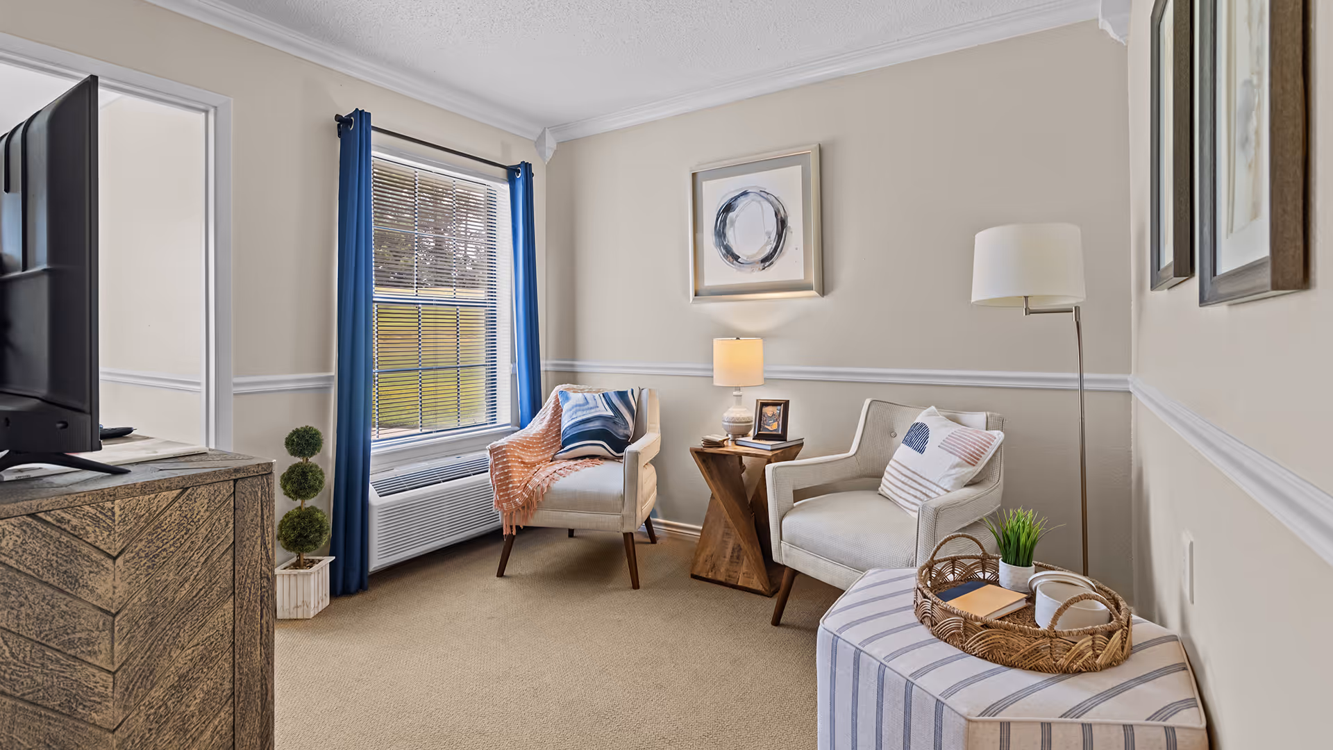 Bright furnished sitting room with two armchairs, a side table and lamp, TV on a console, a window with blue curtains, and decorative accents.