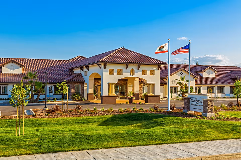 Exterior view of Cedarbrook Memory Care Community building with a tiled roof, arched entrance, two flagpoles displaying the California state flag and the American flag, landscaped lawn, and clear blue sky.