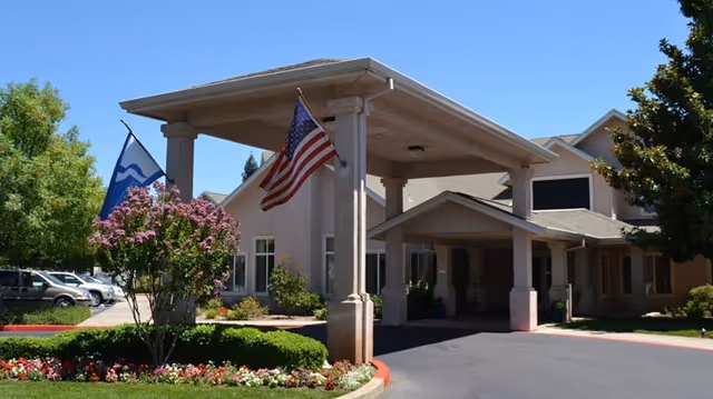 Entrance of a senior living facility with a covered porte-cochere, an American flag, landscaping, and parked cars.