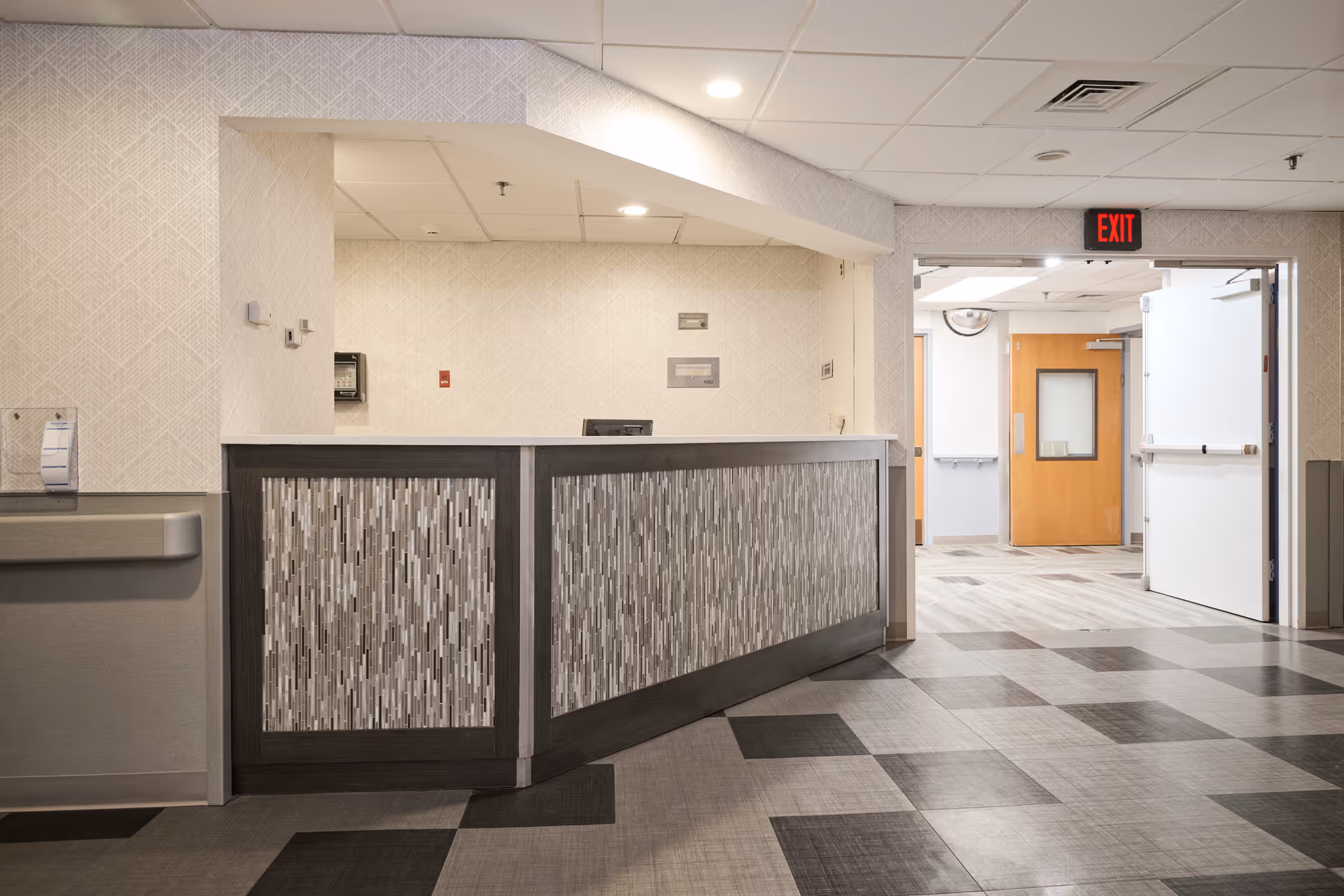 Reception desk area inside a healthcare facility with a patterned front panel, light-colored walls, and a tiled floor with a checkered pattern. There is an open doorway leading to a hallway with wooden doors and an illuminated red exit sign above the door.