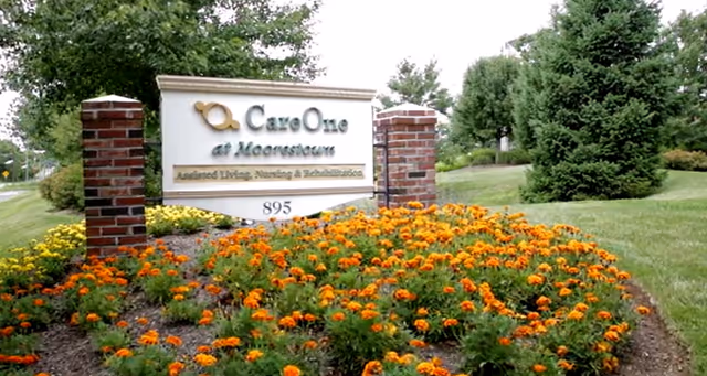 Outdoor view of a CareOne at Moorestown sign surrounded by a flower bed with orange flowers and green grass, with trees and shrubs in the background.