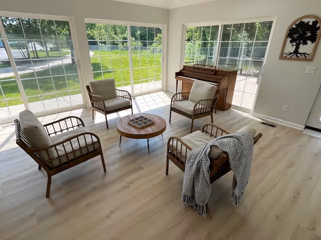 Bright sunroom-style living area with four cushioned wicker chairs arranged around a round coffee table, large windows and a piano by sliding glass doors.