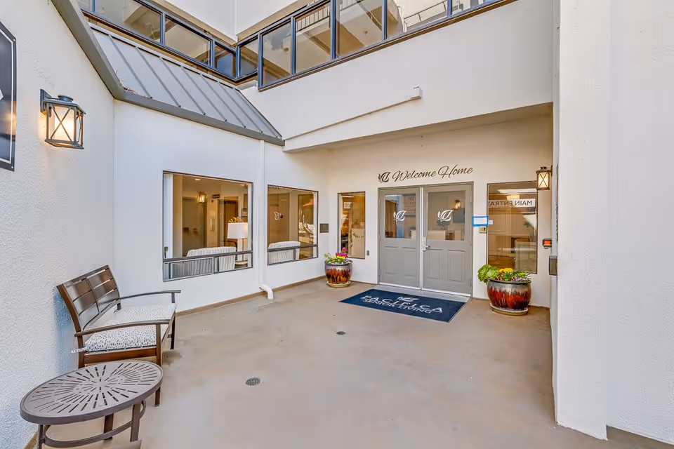 Entrance area of Burlingame Senior Living featuring double gray doors with glass panels, a welcome mat, two large flower pots with plants on either side of the doors, a bench with a small round table on the left, and windows showing a glimpse of the interior seating area. The walls are white, and there are wall-mounted lantern-style lights.