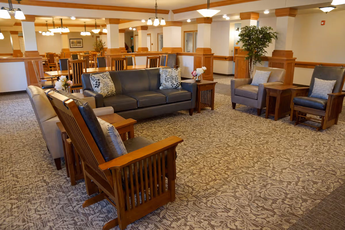 A spacious senior living facility common area with a seating arrangement including a dark leather sofa, wooden armchairs with cushions, and side tables with flower vases. In the background, there are dining tables and chairs under warm ceiling lights, with wooden paneling on the walls and a patterned carpet covering the floor.