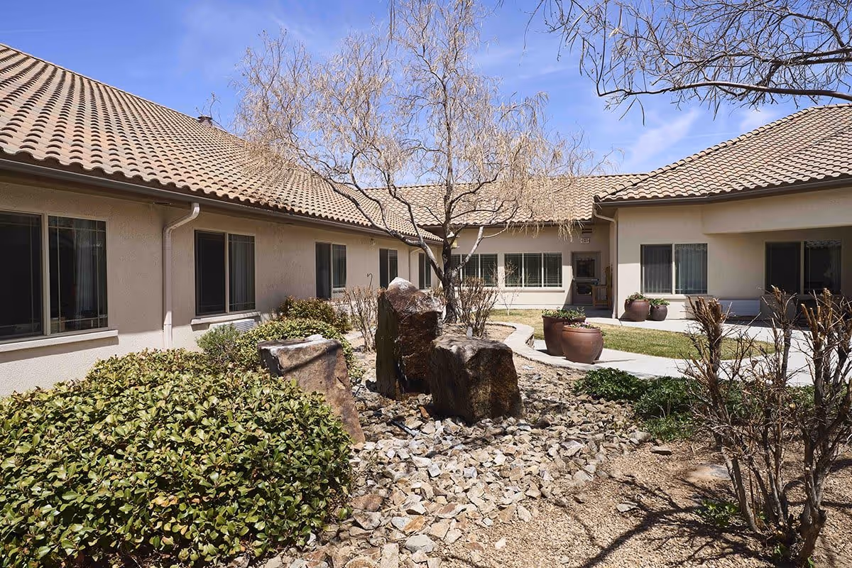 Outdoor courtyard area of a senior living facility with beige stucco buildings and tiled roofs surrounding a landscaped garden featuring rocks, shrubs, a leafless tree, and large potted plants under a clear blue sky.