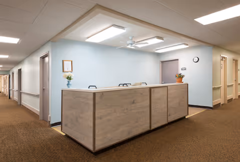 Reception desk area in a nursing and rehabilitation facility with light blue walls, a wooden desk, ceiling lights, a ceiling fan, and a hallway with doors leading to rooms.