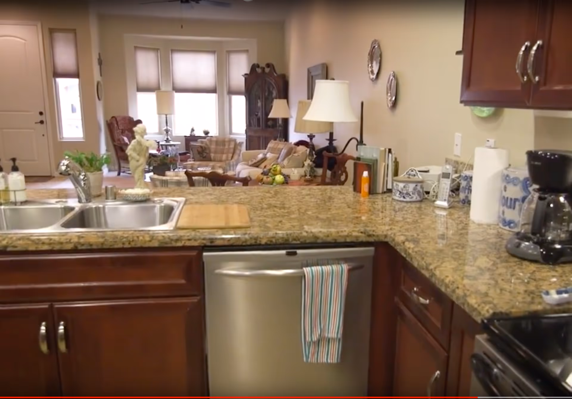 View of a kitchen with granite countertops, a double sink, a dishwasher with a striped towel hanging on the handle, and various kitchen items including a coffee maker and paper towels. In the background, there is a living room area with sofas, armchairs, lamps, and a large window with blinds.