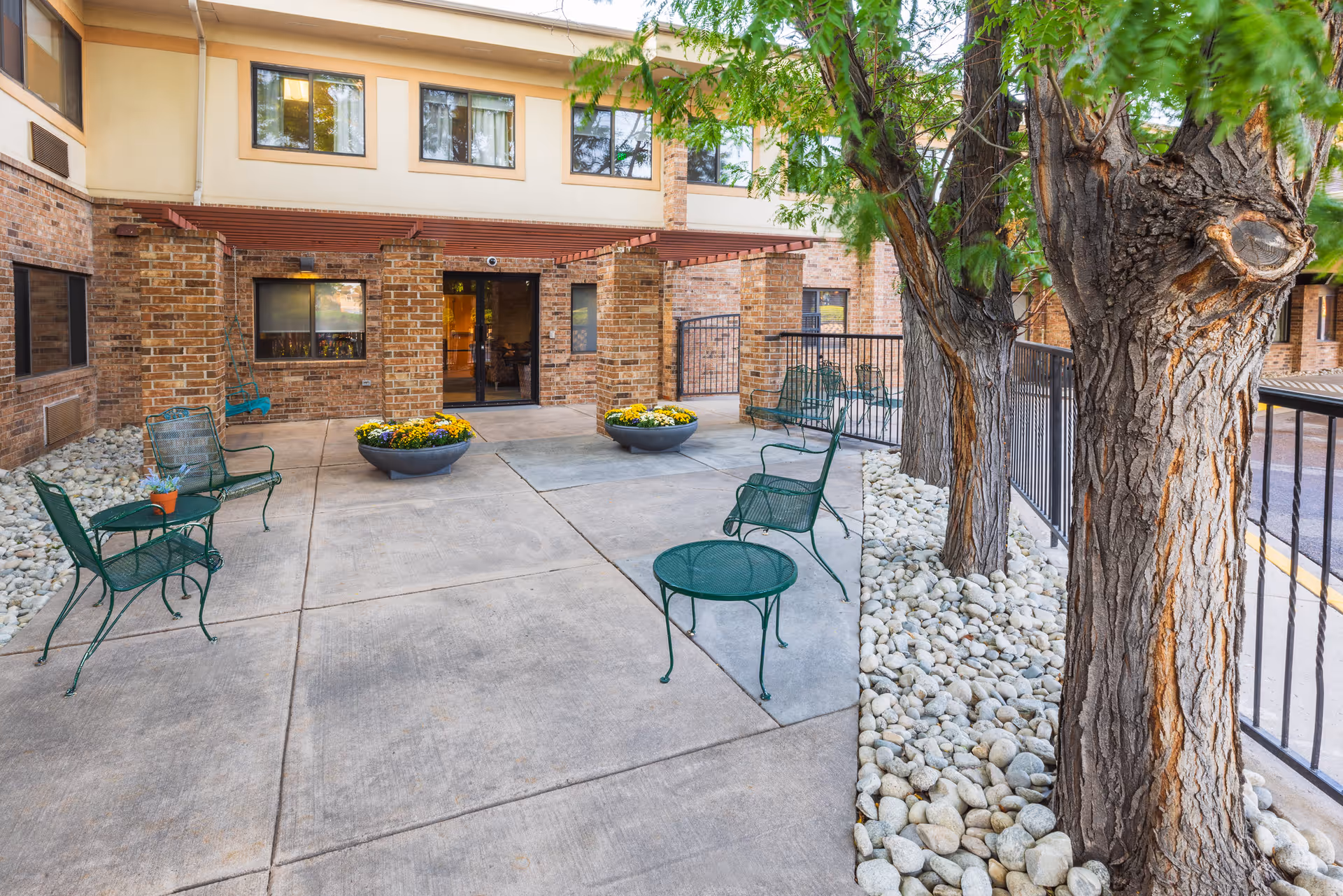 Outdoor patio courtyard with green metal chairs and tables, planter bowls of flowers, trees, and the brick entrance to a senior living building.