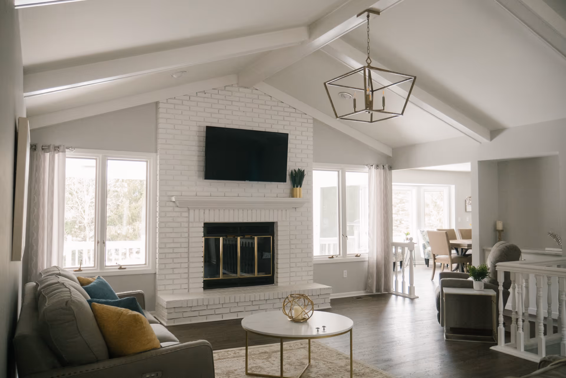 A bright living room with a vaulted ceiling featuring exposed beams, a white brick fireplace with a mounted flat-screen TV above it, a gray sofa with colorful cushions, a round coffee table with decorative items, and large windows with sheer curtains letting in natural light. In the background, a dining area with a table and chairs is visible.