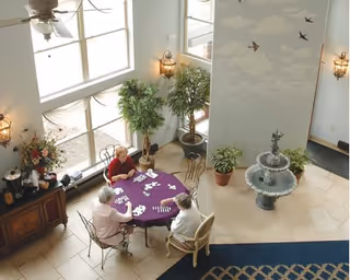 Three elderly women sit around a round table playing dominoes in a bright lobby with large windows, potted plants, and a decorative fountain.