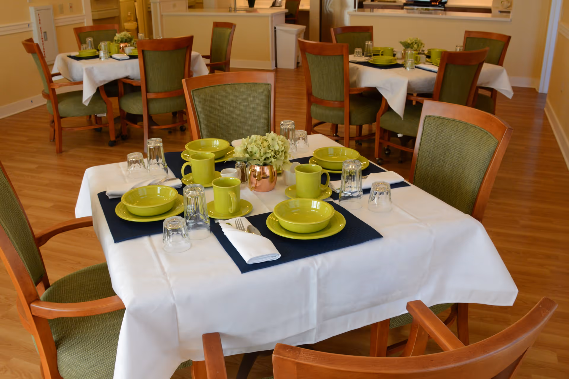 A dining area with wooden chairs featuring green upholstery around tables covered with white tablecloths. Each table is set with green plates, bowls, mugs, silverware wrapped in white napkins, and upside-down drinking glasses. A small floral centerpiece is placed in the middle of the tables. The floor is wooden, and the background shows more similarly arranged tables.