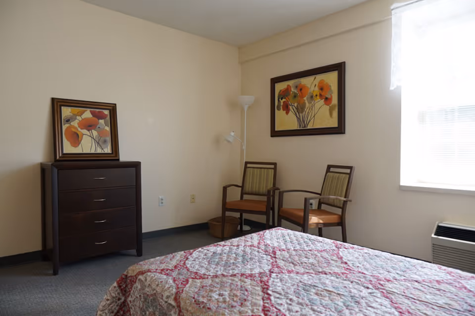 A bedroom with a bed covered in a patterned quilt, two wooden chairs with striped cushions, a dark wooden dresser with a framed floral painting resting on top, a floor lamp, and a window with sheer curtains letting in natural light.
