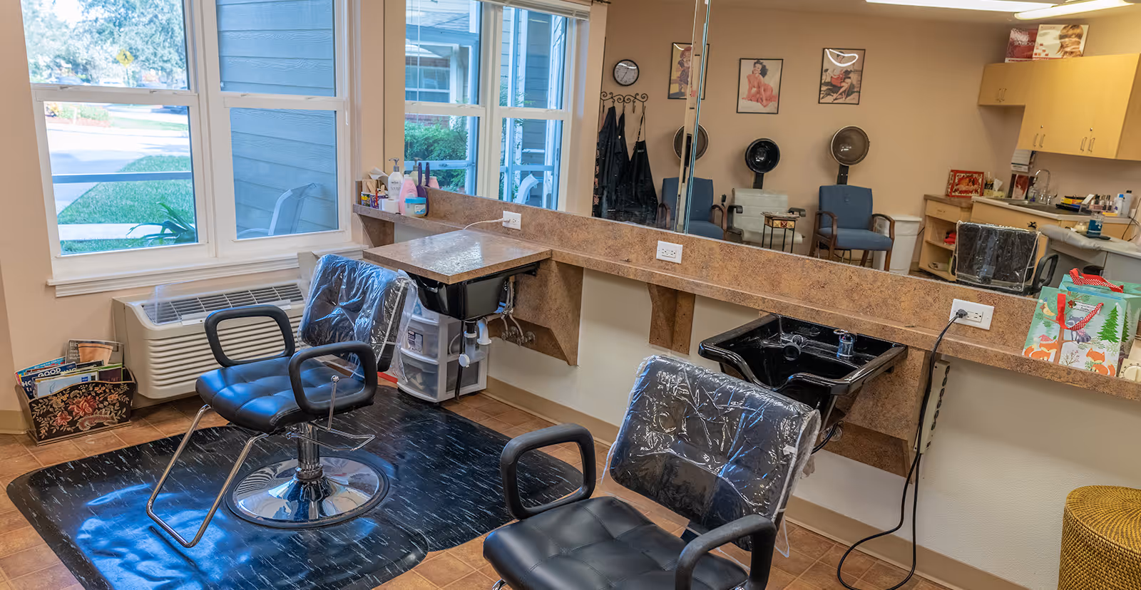 Interior view of a hair salon area in a senior living facility with two black salon chairs covered in plastic, two black hair washing sinks, a large mirror on the wall, a counter with hair care products, and a window showing the outside.