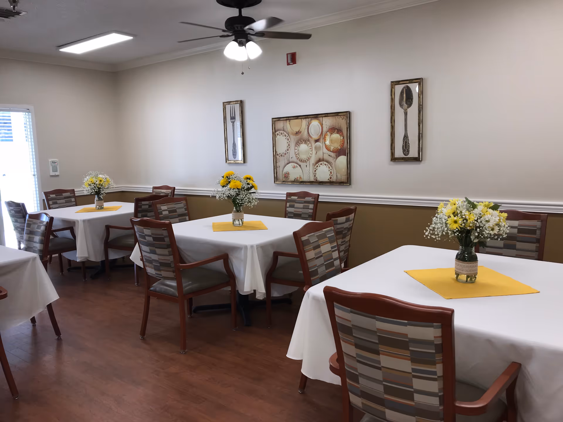 A dining room with several tables covered with white tablecloths and yellow placemats, each table decorated with a vase of yellow and white flowers. The room has wooden flooring, patterned chairs, and wall art featuring utensils. A ceiling fan with lights is visible on the ceiling.