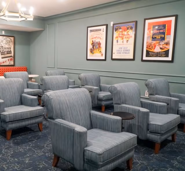 A cozy room with multiple blue upholstered armchairs arranged in rows, each with a small round side table. The walls are painted green and decorated with framed vintage movie posters. A chandelier hangs from the ceiling providing warm lighting.