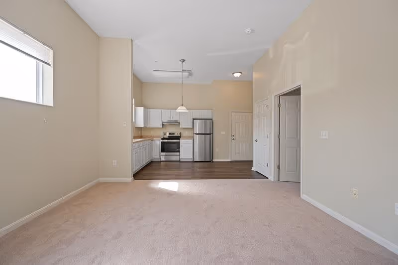 An empty interior space featuring a small kitchen area with white cabinets, stainless steel appliances including a refrigerator and stove, and a hanging light fixture. The kitchen has wood flooring, while the adjacent living area is carpeted. There is a window on the left wall allowing natural light, and two doors on the right side of the image.