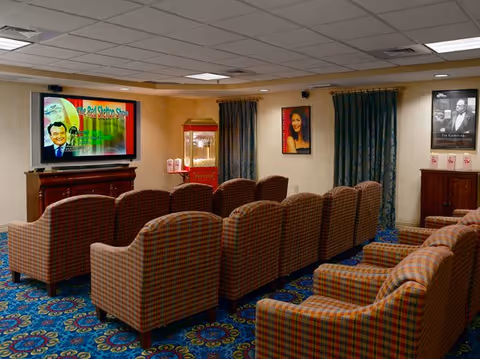 A cozy media room with two rows of plaid upholstered armchairs facing a large flat-screen TV mounted on the wall. The TV screen displays a colorful image titled 'The Red Skelton Show.' There is a popcorn machine in the corner, framed pictures on the walls, and blue patterned carpet covering the floor.