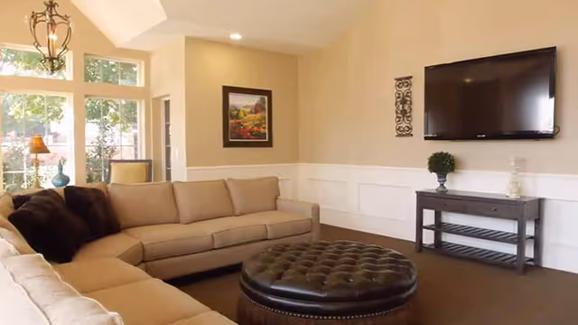 A cozy living room with a large beige sectional sofa, a round tufted leather ottoman in the center, a wall-mounted flat-screen TV above a dark wooden console table, and a framed painting on the wall. The room has beige walls with white wainscoting and large windows letting in natural light.