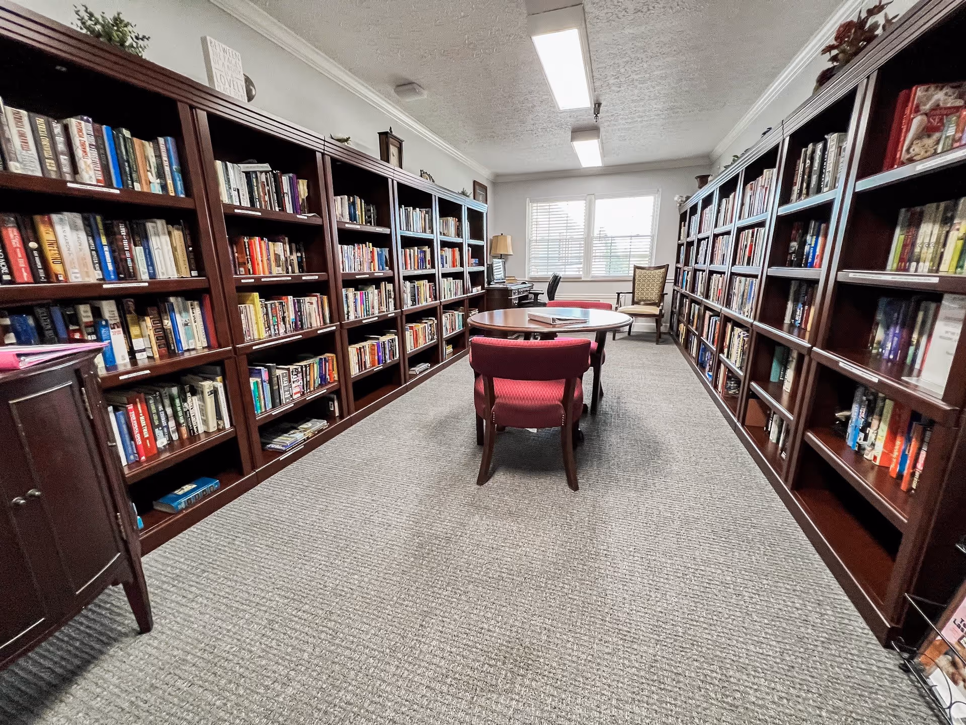 A quiet library room with tall wooden bookshelves filled with books lining both sides of the room. In the center, there is a round wooden table with four red upholstered chairs around it. At the far end near the window, there is a desk with a computer and a lamp, and a single wooden chair with a cushion. The room is carpeted and well-lit with fluorescent ceiling lights.