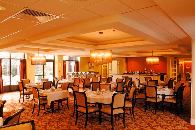 A warmly lit dining room with multiple round and rectangular tables covered with white tablecloths, set with glassware and silverware. The room features patterned carpet, upholstered chairs, large windows with curtains, and hanging pendant lights. There is a buffet area in the background.