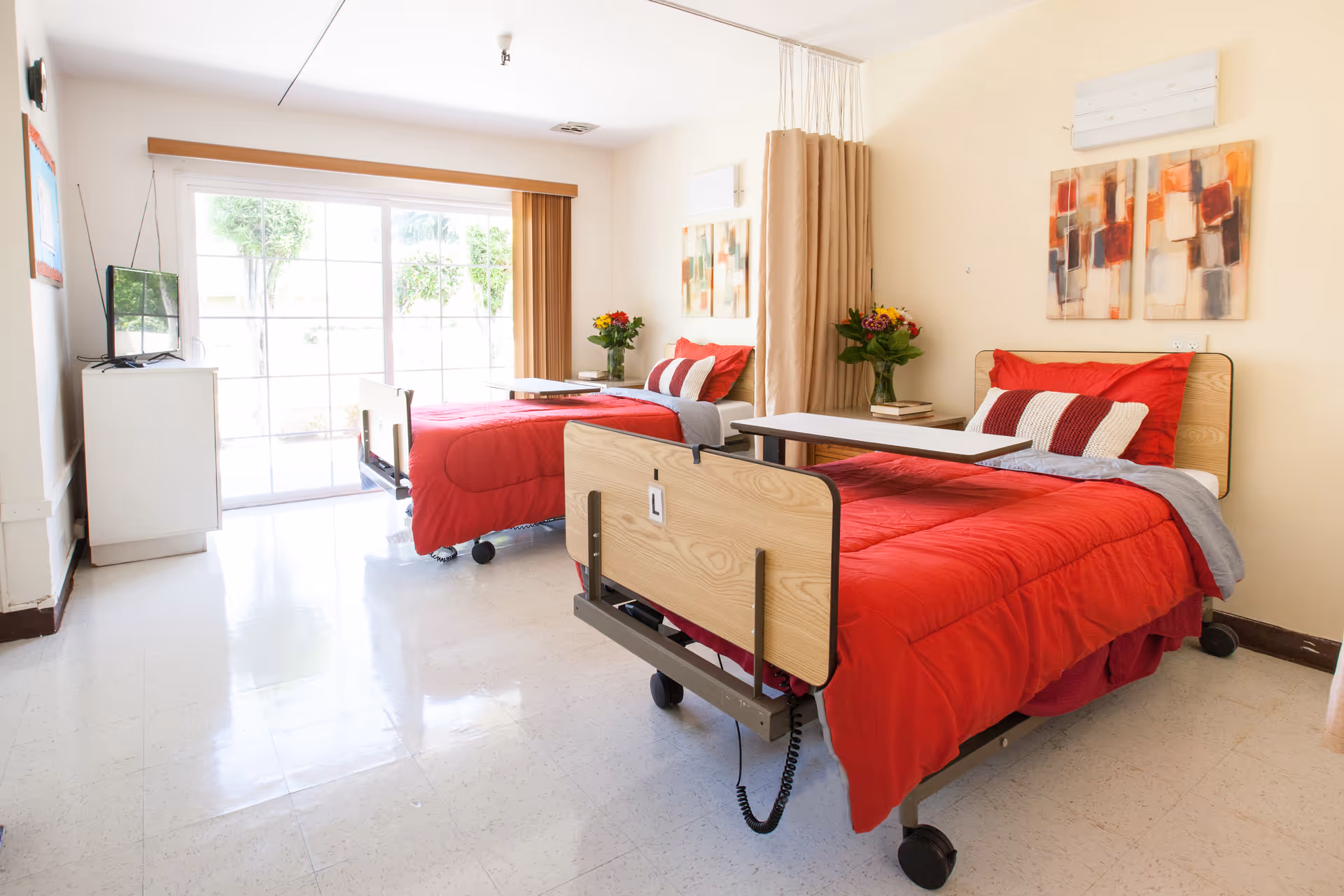 A bright and clean room with two hospital-style beds, each made with red bedding and pillows. Each bed has an overbed table and a bouquet of flowers on a small side table. There is a large sliding glass door letting in natural light, a small TV on a white cabinet, and abstract artwork on the walls.