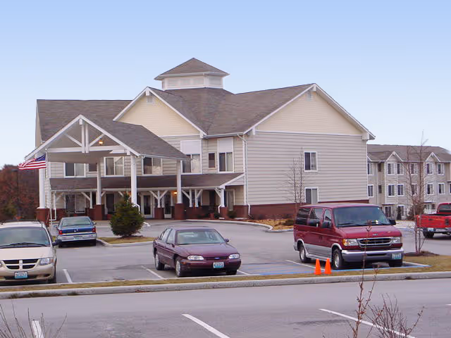 Exterior view of St. Andrew's Apartments at Kirksville, showing a multi-story building with beige siding and a covered entrance. Several cars are parked in the parking lot in front of the building, and an American flag is visible near the entrance.