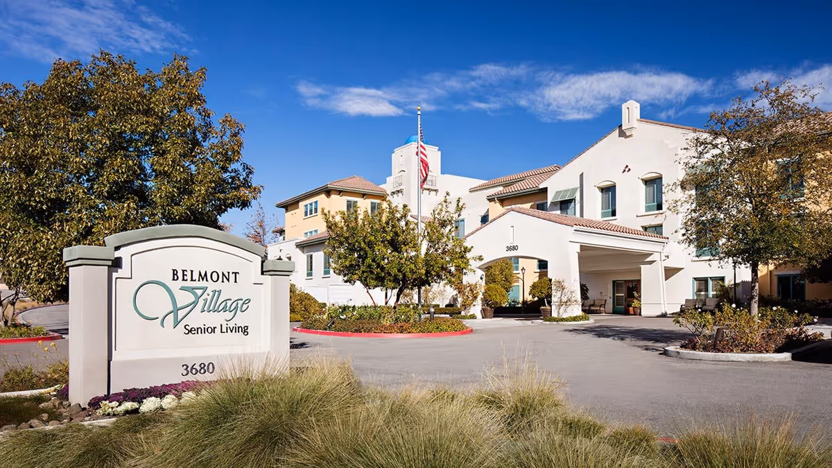 Exterior view of Belmont Village Senior Living facility with a clear blue sky, trees, and a sign displaying the facility name and address 3680. The building has a light-colored facade with multiple windows and a covered entrance.