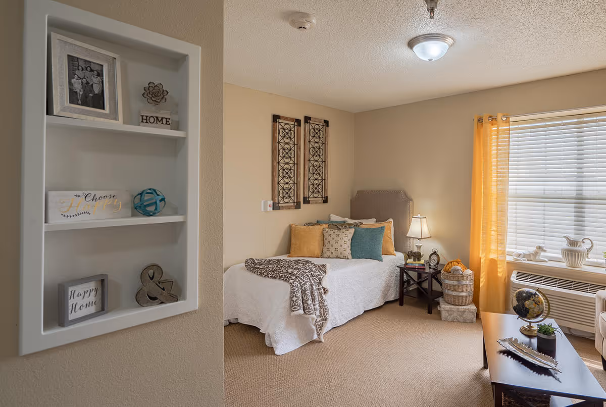 A cozy bedroom in Mackenzie Court featuring a single bed with multiple decorative pillows and a patterned throw blanket. The room has beige walls, a window with yellow curtains, and a small side table with a lamp and decorative items. A built-in wall shelf near the entrance holds framed photos and decorative pieces. There is a coffee table with a globe and small plants, and the room is carpeted.
