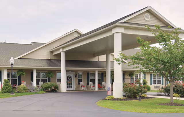 Exterior view of a senior living facility named Amity Place showing the front entrance with a covered drop-off area supported by columns, surrounded by landscaped greenery and a paved driveway.