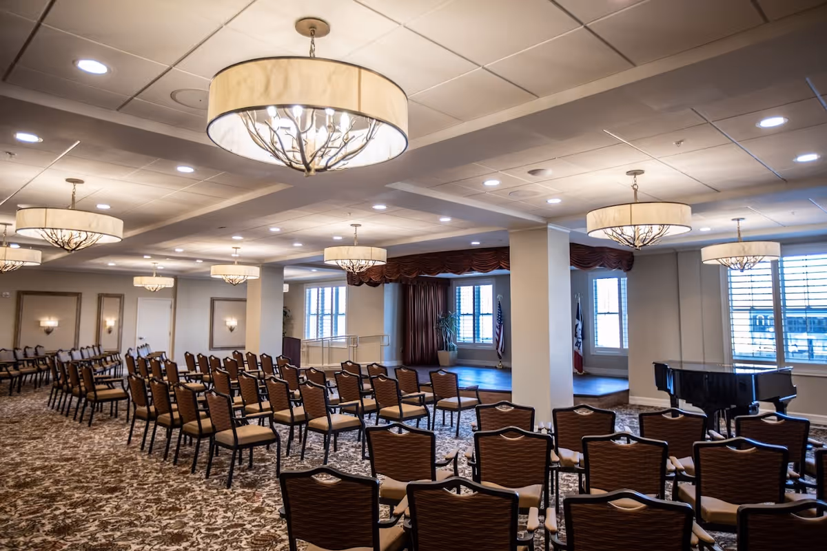 A large, well-lit event room with rows of chairs arranged facing a small stage area. The room features elegant chandeliers, patterned carpet, and large windows with blinds. A black grand piano is positioned near the windows on the right side of the room.