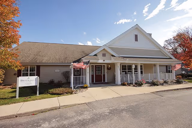 Exterior view of a single-story building with beige siding and a gabled roof, featuring a covered porch with white railings and two American flags on either side of the entrance. There is a sign on the lawn that reads 'Brookdale Midland'. Trees with autumn foliage are visible on both sides of the building.