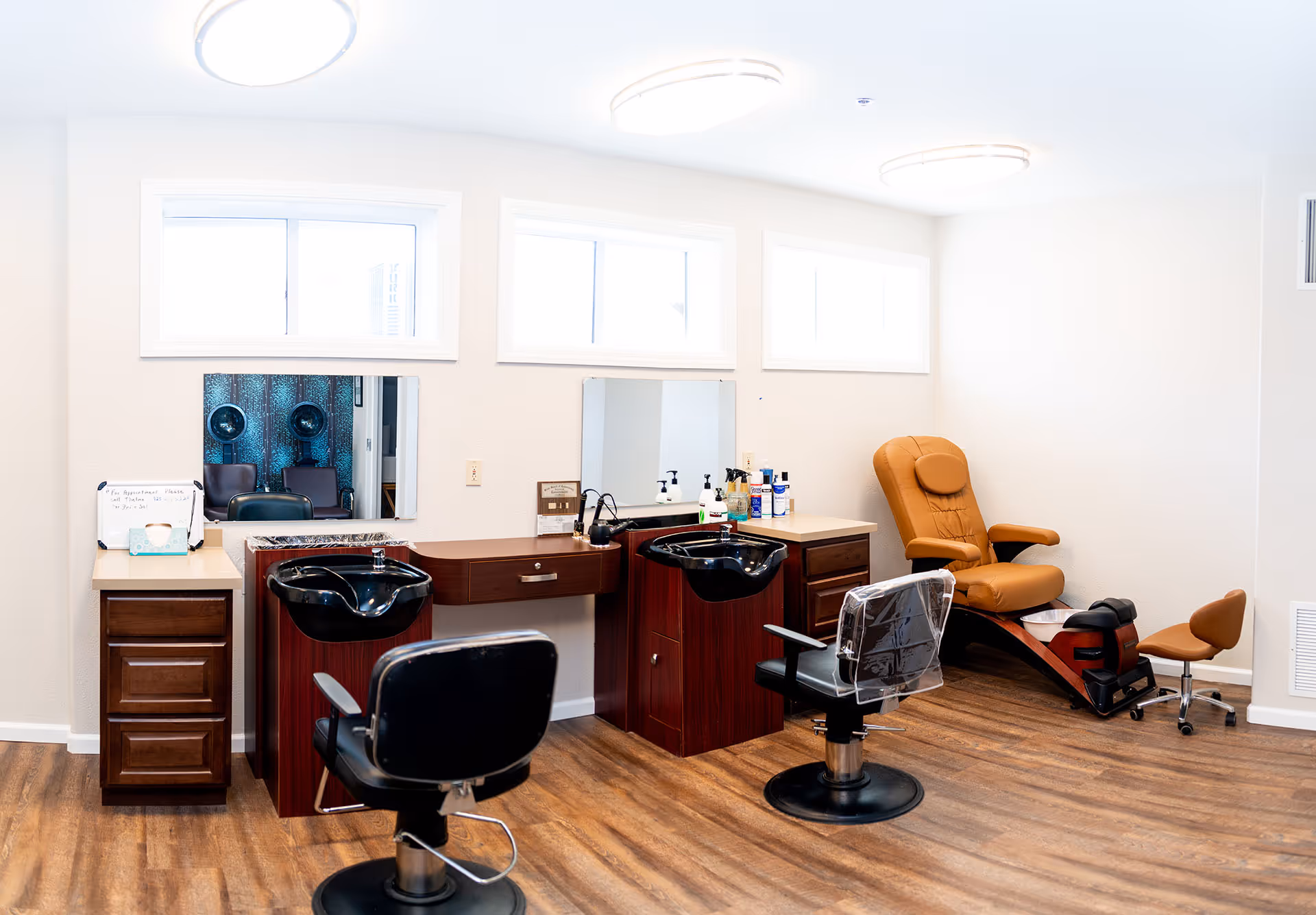 A bright and clean salon area in a senior living facility featuring two black salon chairs in front of sinks and mirrors, a tan reclining pedicure chair with a footrest, and wooden flooring. The walls are white with three rectangular windows near the ceiling allowing natural light in.