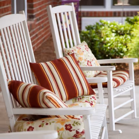 White wooden rocking chairs with striped and floral cushions arranged on a covered porch with a brick wall and green shrubbery in the background.