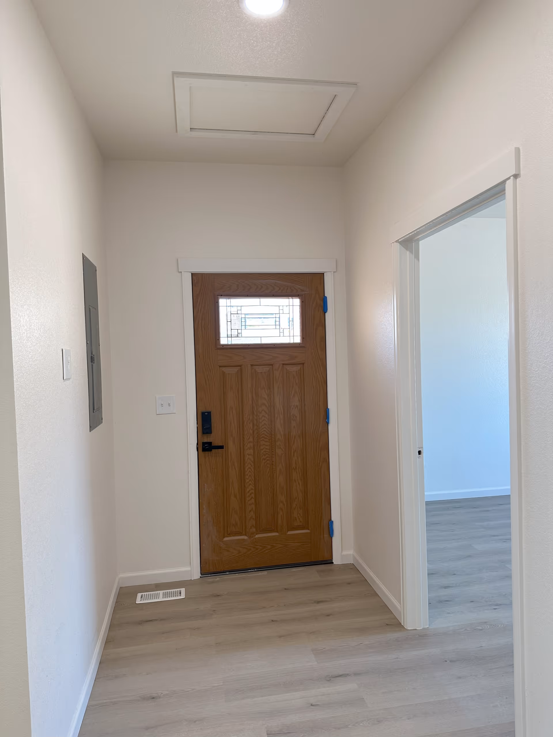 Interior view of a hallway with a wooden front door featuring a decorative glass window. The hallway has light-colored walls and wood flooring. There is a vent on the floor near the door and an electrical panel on the left wall. An open doorway on the right leads to another room with similar flooring and light walls.