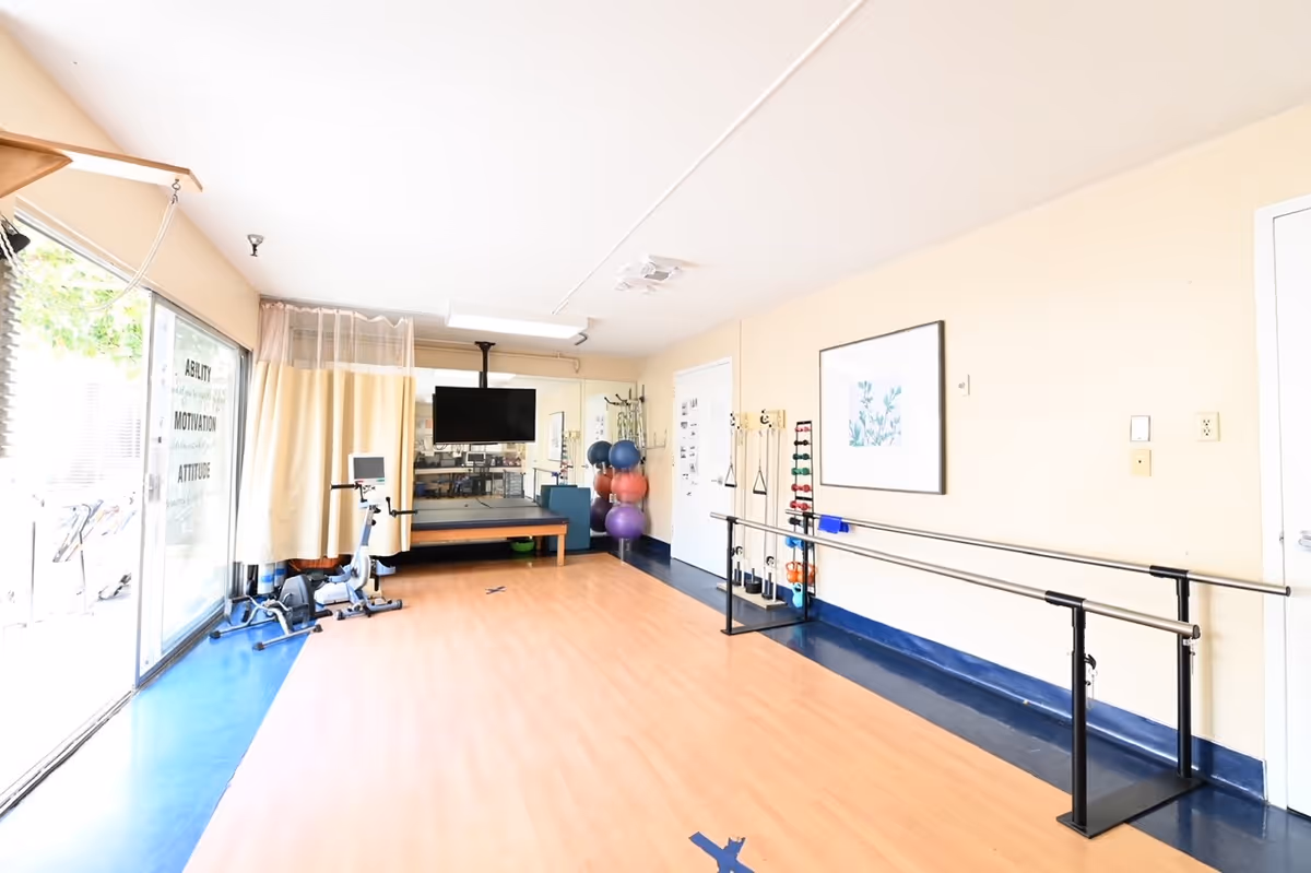 A bright rehabilitation or exercise room with wooden flooring and blue trim. The room contains parallel bars for walking practice, exercise balls stacked in a corner, a stationary bike, a TV mounted on the wall, and a rack with various weights. Large windows with sheer curtains allow natural light to enter, and motivational words like 'ABILITY', 'MOTIVATION', and 'ATTITUDE' are visible on the glass door.