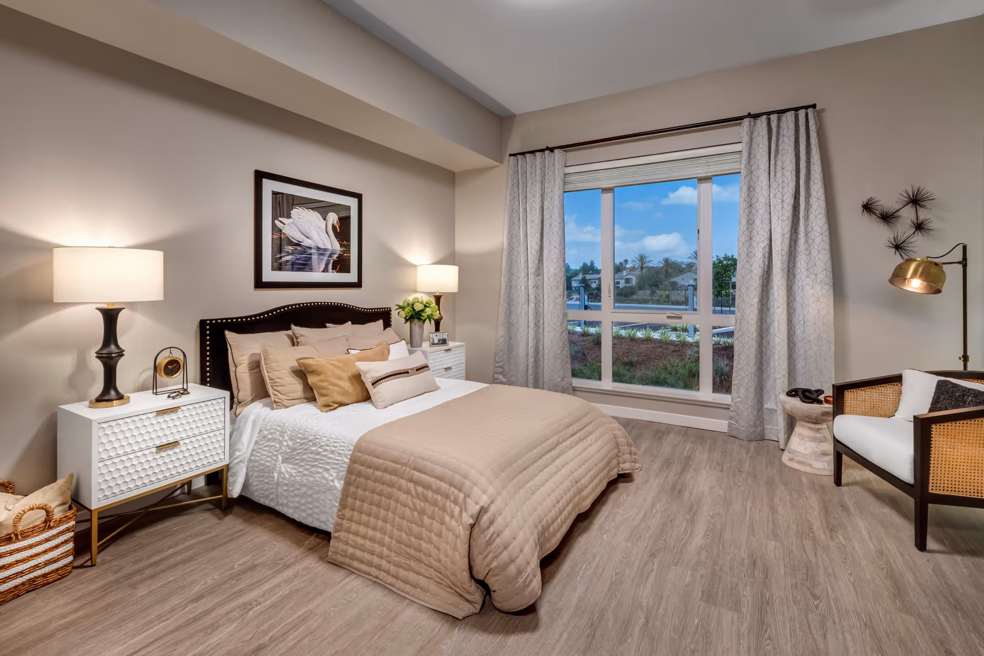 A cozy bedroom with a neatly made bed featuring beige and white bedding and multiple pillows. Two bedside tables with lamps flank the bed. A framed picture of a swan hangs above the headboard. A large window with curtains shows an outdoor view with greenery and houses. A cushioned chair and a small side table with a lamp are positioned near the window. The room has wood flooring and neutral-colored walls.