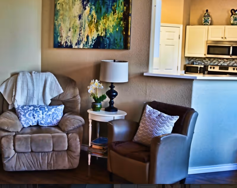 A cozy living area with a beige recliner chair draped with a white blanket and a blue patterned pillow, next to a small white side table holding a black lamp and a potted orchid. Adjacent is a brown armchair with a white decorative pillow. In the background, a kitchen area with white cabinets, a microwave, and a stove is visible, along with a colorful abstract painting on the wall.