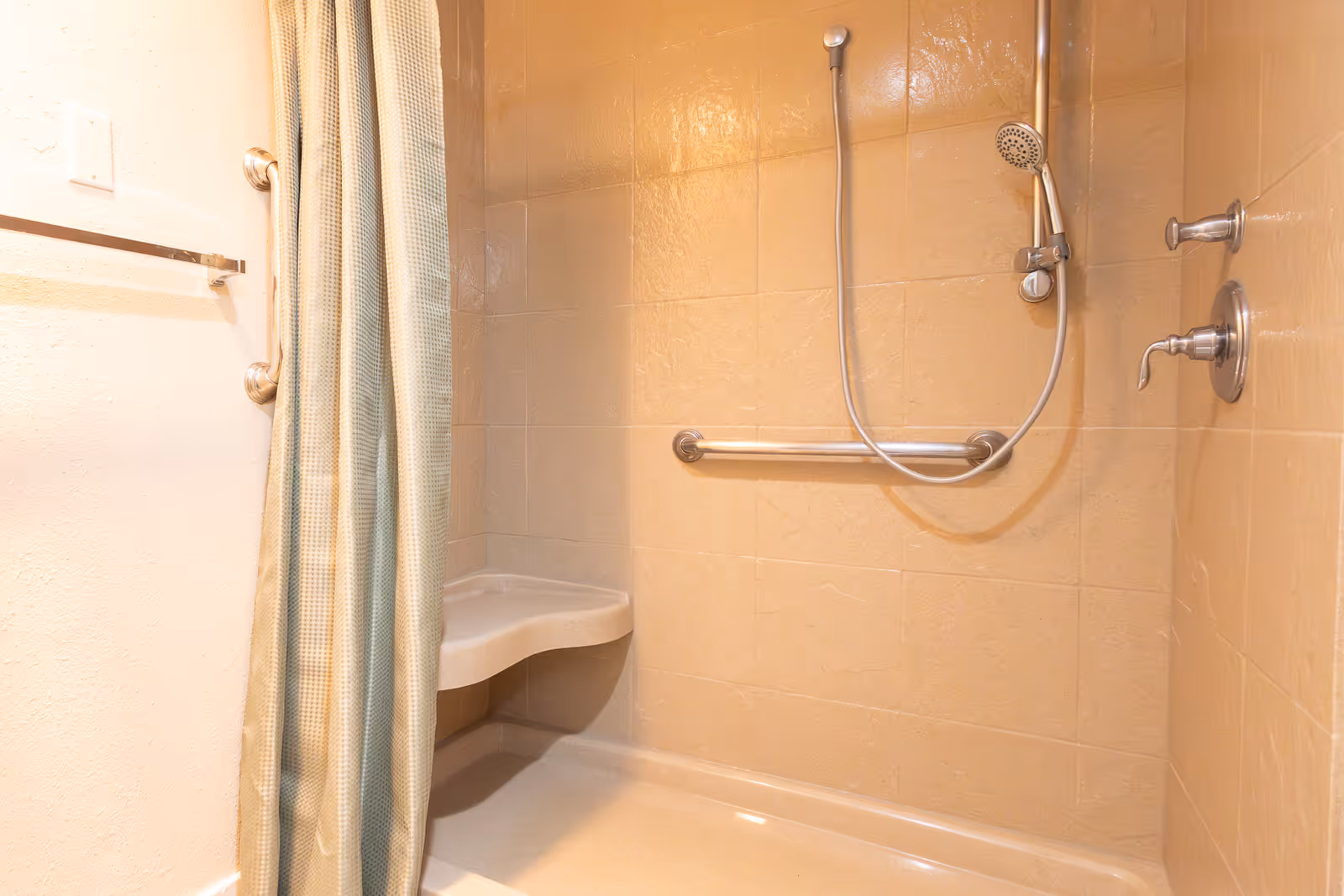 Shower area with beige tiled walls, a handheld showerhead, a built-in corner seat, and grab bars. A shower curtain is partially visible on the left side.