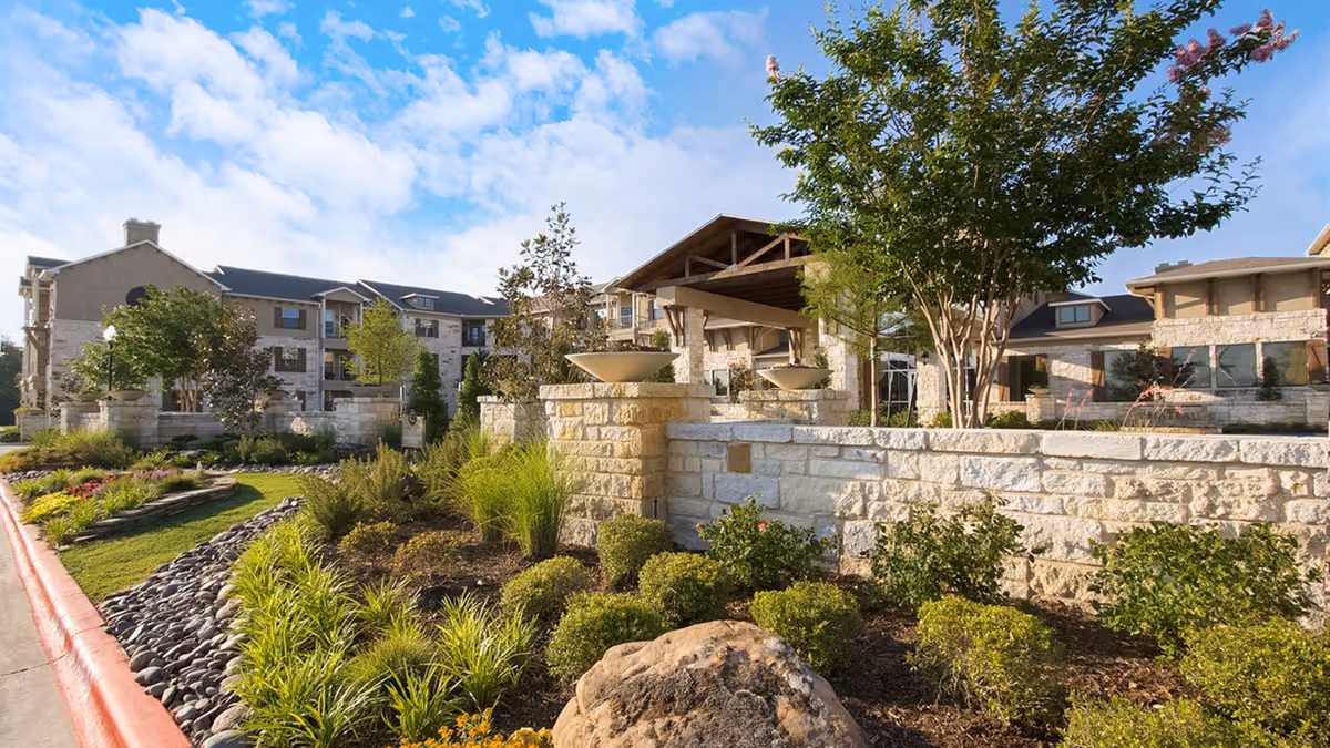 Exterior view of Chateau McKinney senior living facility showing stone walls, landscaped garden with shrubs and trees, and a multi-story building under a partly cloudy blue sky.