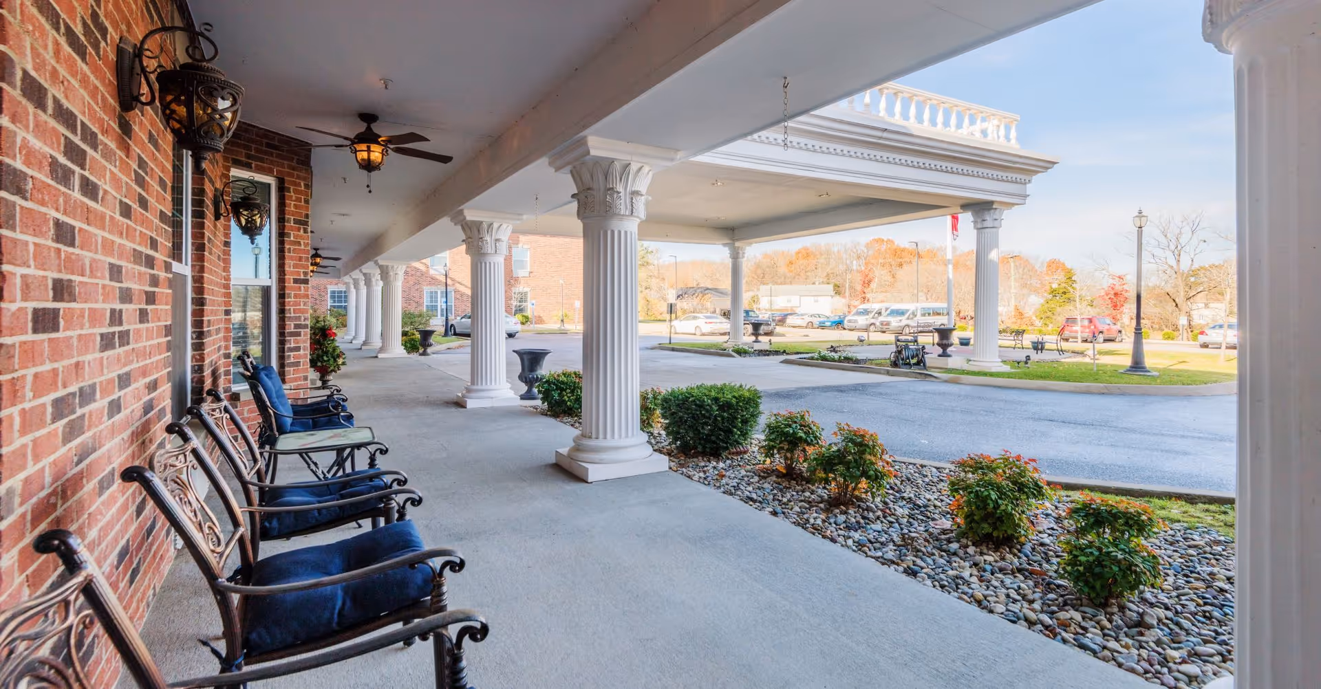Covered outdoor seating area with several metal chairs with blue cushions along a brick wall. White decorative columns support the roof, and ceiling fans with lights are mounted overhead. In the background, there is a driveway, some bushes, and parked cars under a clear sky.